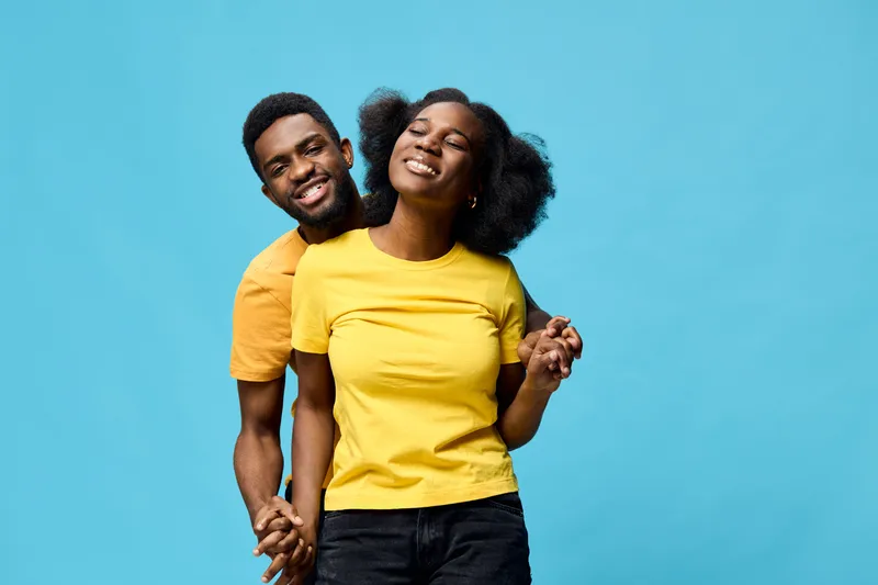 Young African couple wearing bright yellow t shirts, smiling and holding hands against a vibrant blue background, representing love and happiness