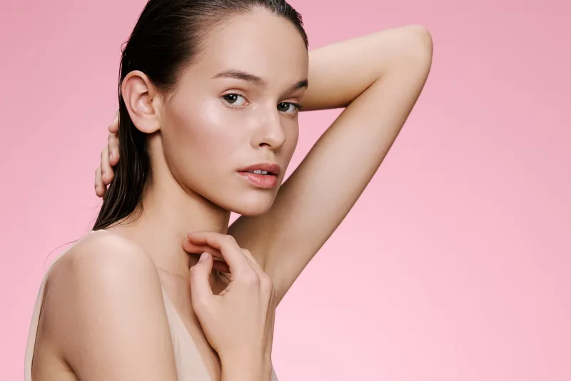 Young female model with natural beauty, minimal makeup, and wet hair posing against a soft pink background, conveying confidence and simplicity
