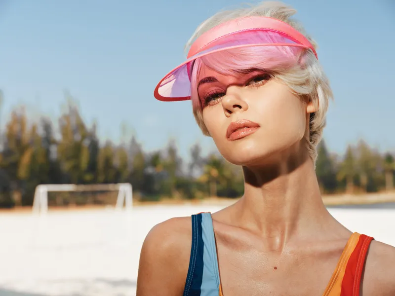 Portrait of a stylish woman wearing a pink visor on a sunny beach, sunlit skin and vibrant makeup, conveying a bright, confident summer vibe and fashionable allure.