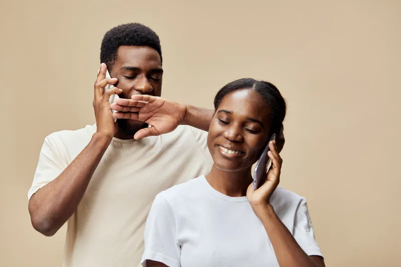 Young black couple communicating joyfully on smartphones against a warm beige background, showcasing technology and connection