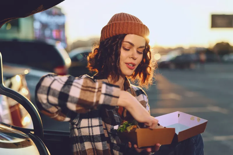 Woman with takeaway food by car trunk in outdoor evening light, casual beanie style and authentic candid lifestyle moment with golden hour glow, mindful living and emotional storytelling vibe.
