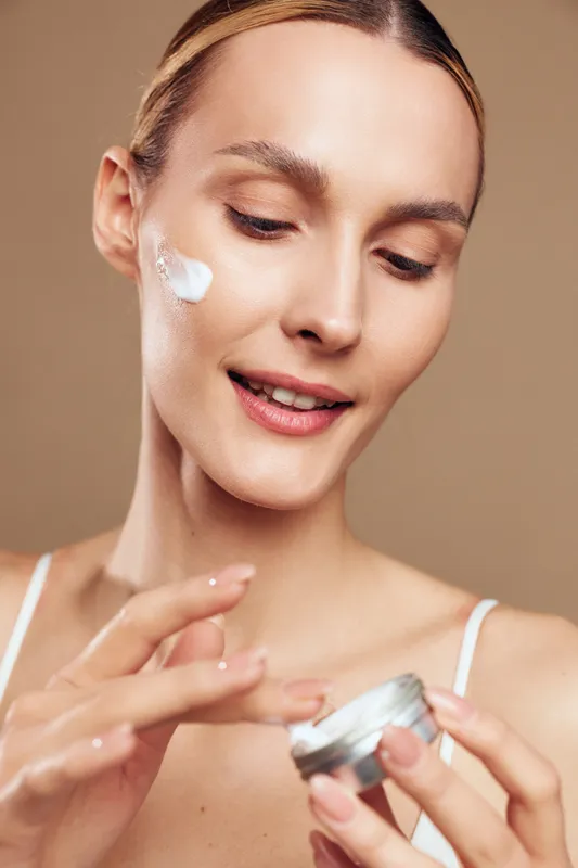Bright, focused portrait of a woman applying moisturizer on her cheek, soft lighting and close-up view, calm mood, skincare routine highlight, natural skin tone