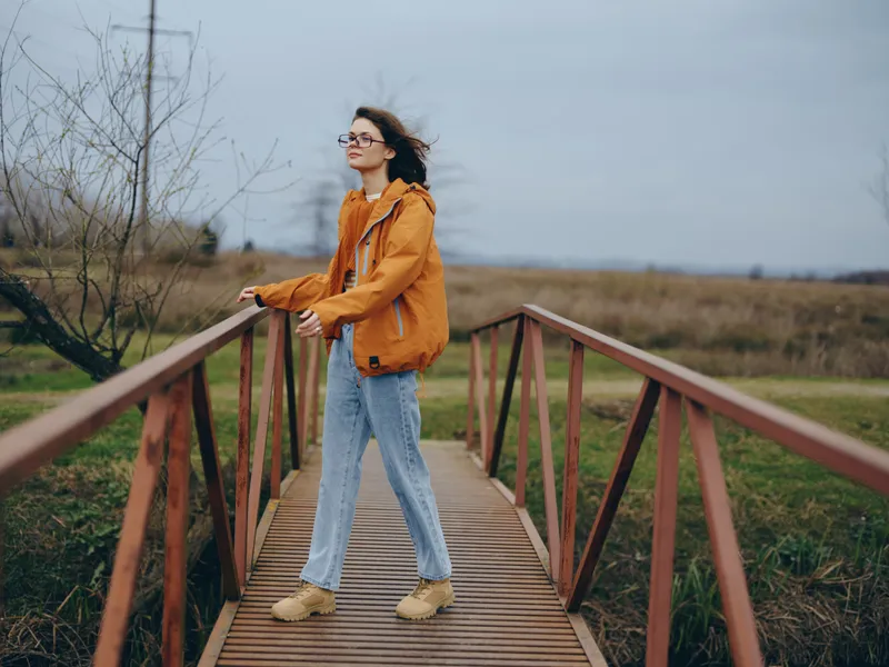 Woman lifestyle portrait on an outdoor wooden bridge, wearing orange jacket and jeans, walking forward in wind, glasses and scarf, countryside field background, calm spring morning