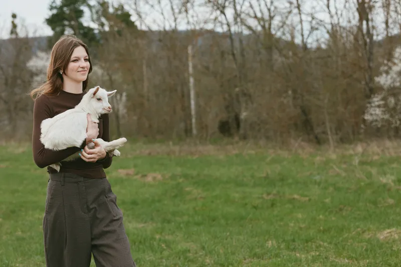 A woman outdoors holds a white goat kid in her arms, smiling softly in a grassy field, depicting a casual lifestyle scene, gentle care, companionship, and farm life.