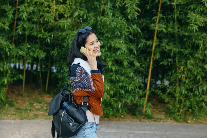 Woman on phone outdoors with backpack, casual jacket and denim, standing by a green hedge, smiling while talking, urban nature setting, everyday communication, street style, carefree moment