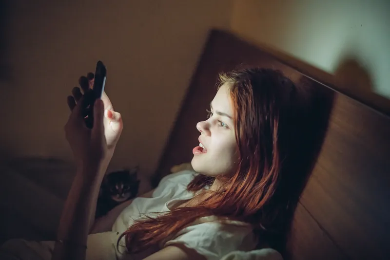Рыжеволосая девушка лежит в постели со смартфоном ночью