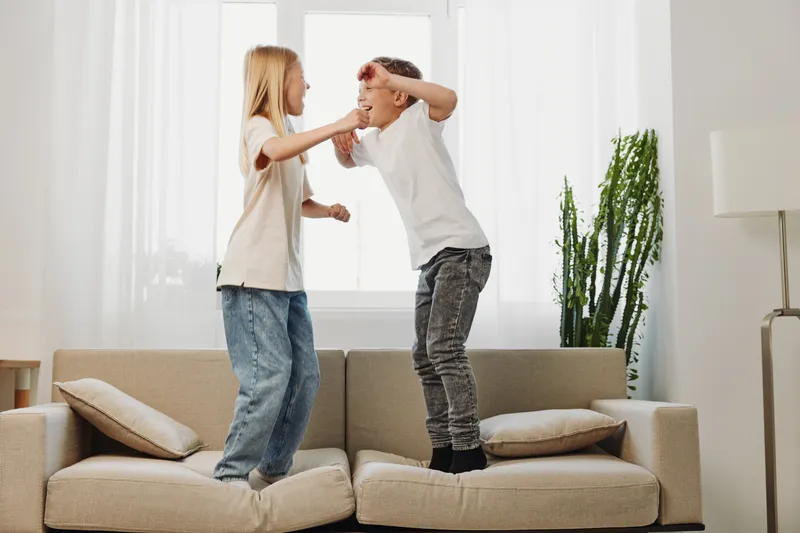 Two children playfully jump on a sofa in a bright living room with large windows and indoor plants. The scene captures joy, energy, and sibling interaction in a modern home.