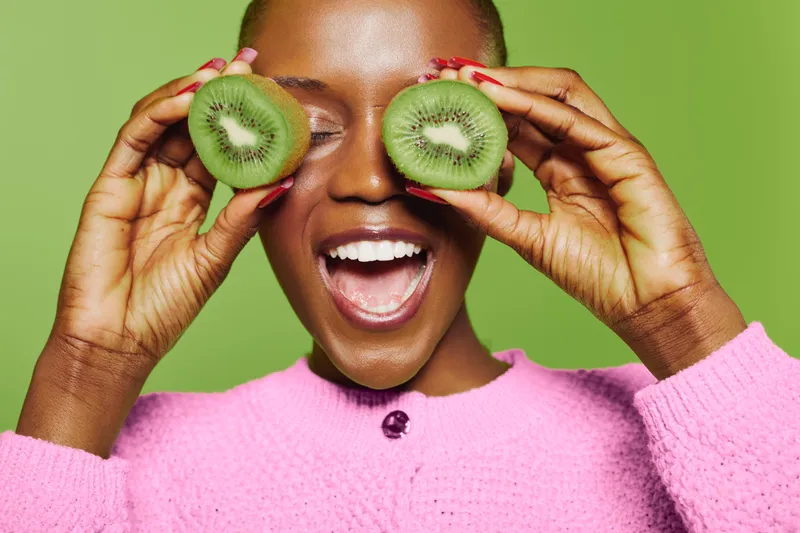 Happy black woman with dark skin holds two kiwi halves over her eyes, smiling with open mouth. Concept of fresh fruit, healthy lifestyle, natural beauty, and skincare in studio on green background.