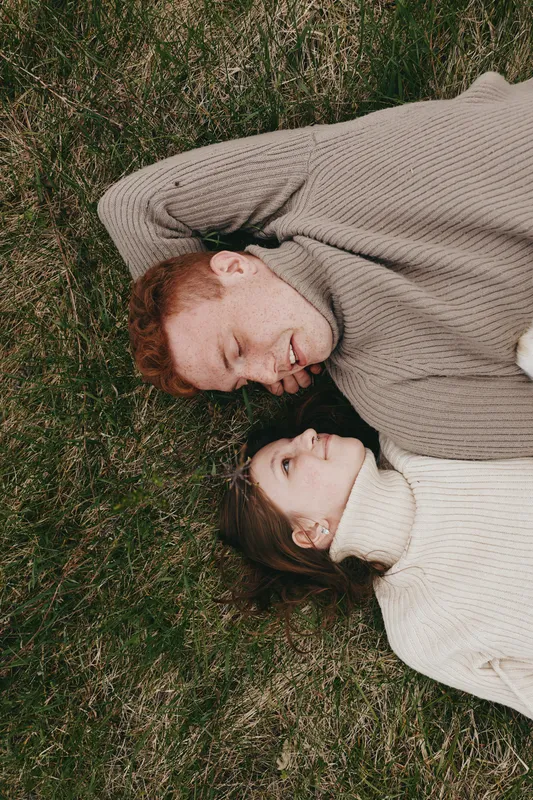 Two people lie side by side on green grass, smiling softly while wearing cozy sweaters in an outdoor moment, warm light, relaxed posture, intimate connection, natural tones, simple countryside vibe,