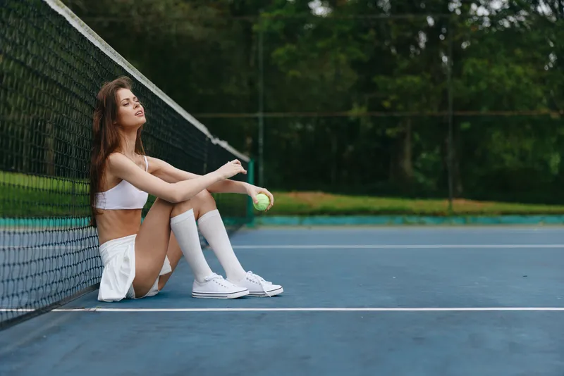 Young sporty woman sitting on a tennis court, enjoying a moment of relaxation while holding a tennis ball, dressed in a stylish tennis outfit