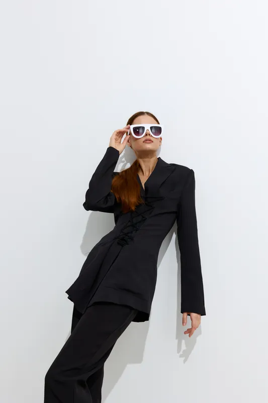 Fashion woman in black stylish outfit wearing white sunglasses posing against white background in minimalistic studio setting with confident and modern look.