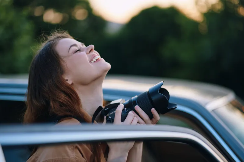 Young smiling woman holding a camera, capturing a moment outdoors, with a joyful expression in a natural setting during golden hour.