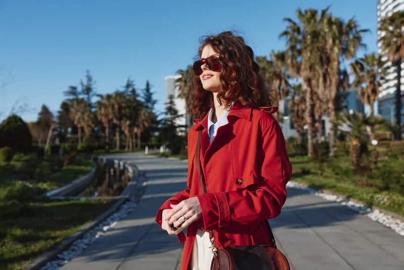 A stylish woman in a red coat walks along a sunny promenade, enjoying warm weather and a relaxed mood.
