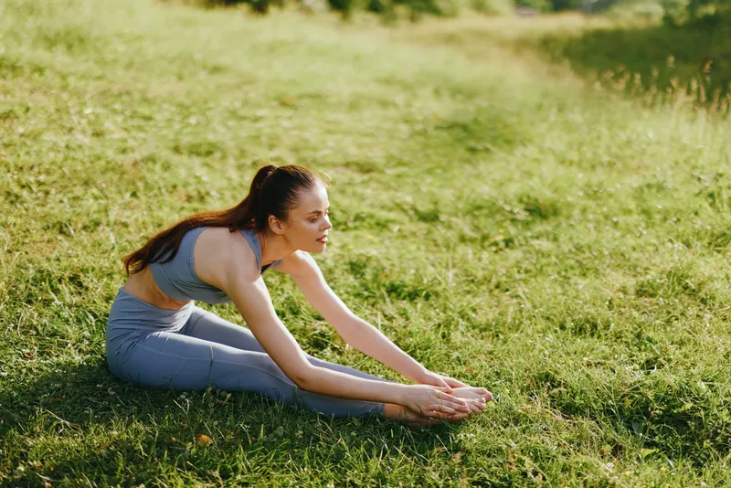 Young woman practicing yoga outdoors on green grass, stretching her body in a peaceful nature setting with bright sunlight, representing wellness and mindfulness