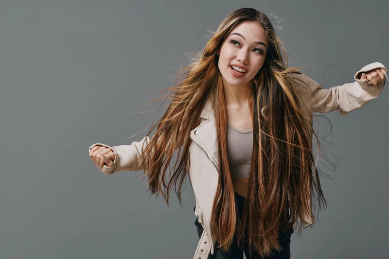 Young woman with long hair and braids wearing beige jacket dancing and smiling against gray background for fashion, beauty, and lifestyle concept.