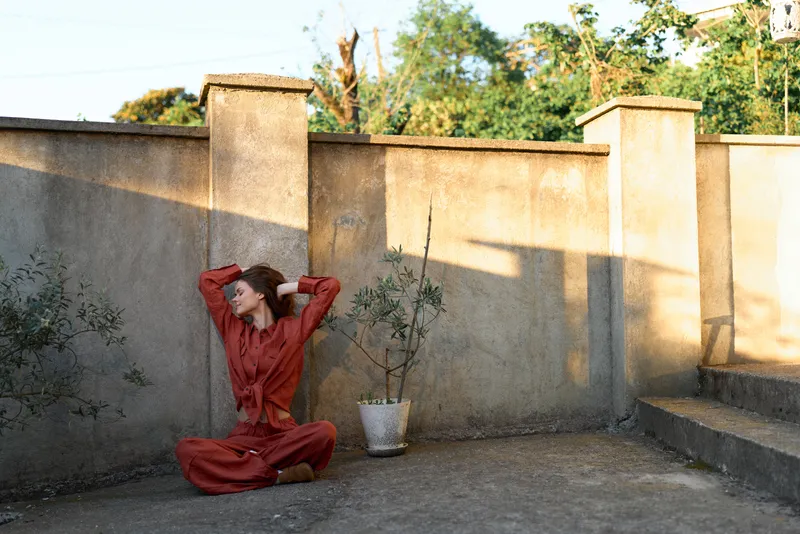 Woman in a flowing red dress sits on a rooftop patio, posing with relaxed mood, leaning against a sunlit wall beside a potted plant, capturing casual outdoor lifestyle