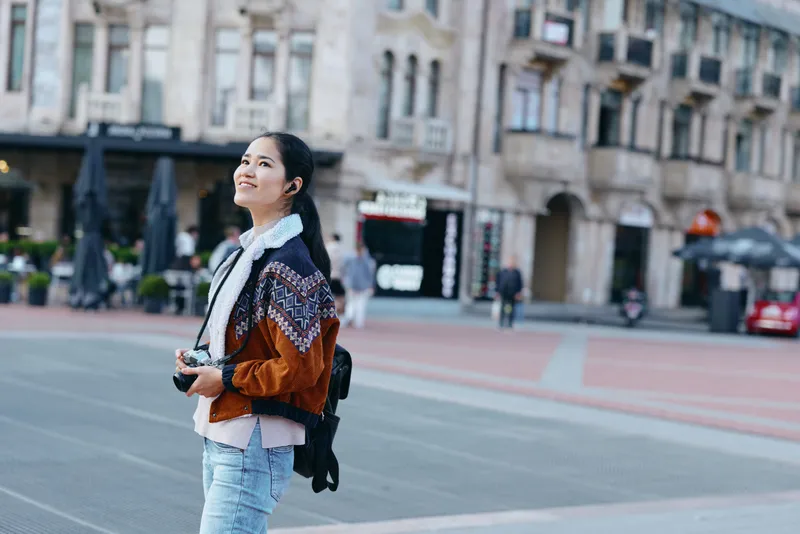 Woman with camera stands on a city street, wearing a patterned jacket and backpack, capturing urban life, architecture and candid moments with a curious gaze.