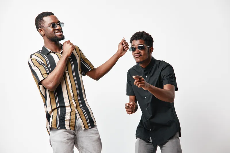 Two young black men with dark skin wearing sunglasses dancing and having fun together on white background. Casual clothing and joyful expressions in studio setting.