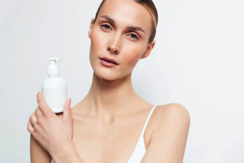 A young woman in a white top holds a pump bottle in a studio, presenting a skincare product with a calm, confident expression against a neutral background and soft lighting.
