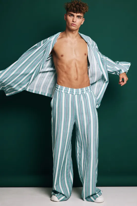 Young man posing in striped loose pants and open shirt with bare chest against dark background. Fashion model with casual style and confident expression in studio photo shoot.