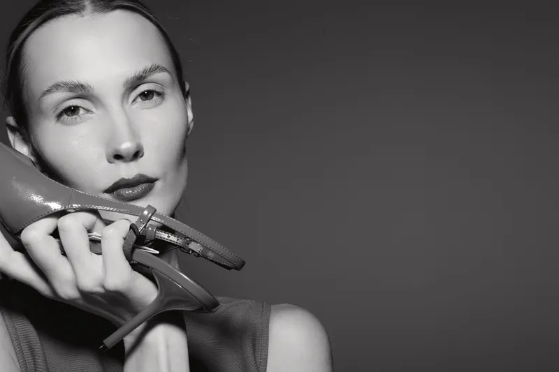 White woman with pale skin holds a high heel red shoe near her face in a minimalist studio with dark background. Concept of fashion and elegance.