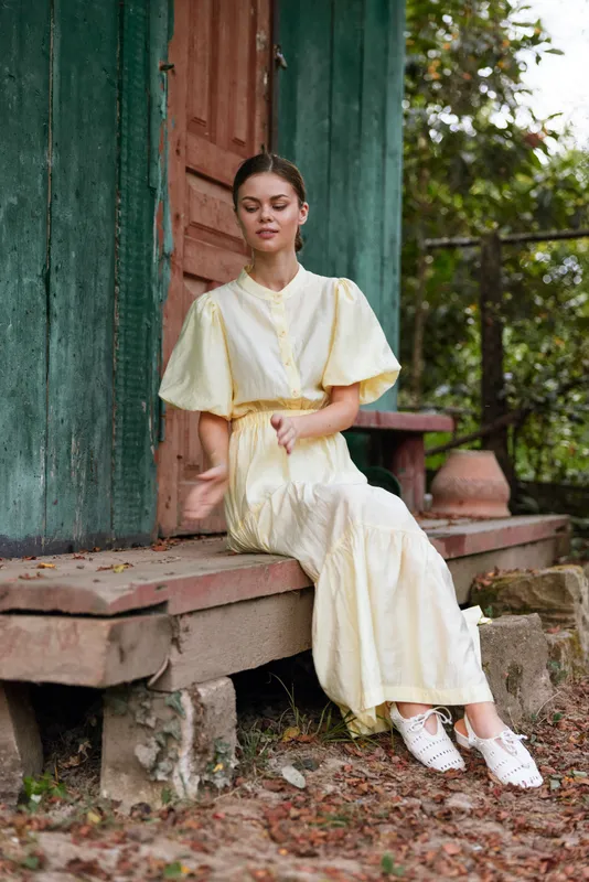 Woman sitting on wooden steps wearing a pale yellow dress in an outdoor porch setting, calm expression, soft natural light, relaxed pose, fashion and serenity.