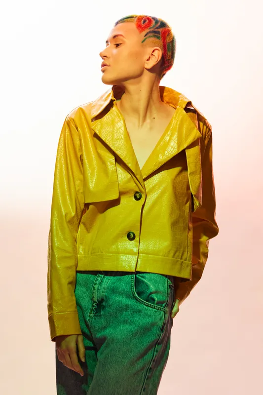 Young woman with colorful braided hair wearing a shiny yellow jacket and green jeans standing against bright white background in a modern fashion style.