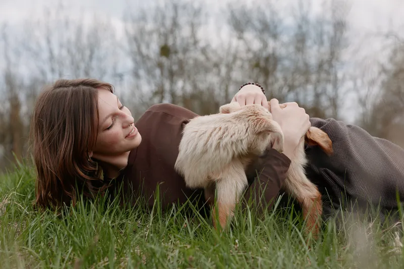 Lying woman outdoors, gentle interaction with a curious goat kid, soft grass underfoot, calm connection, natural colors, tranquil meadow mood, candid lifestyle moment outdoor warmth gently