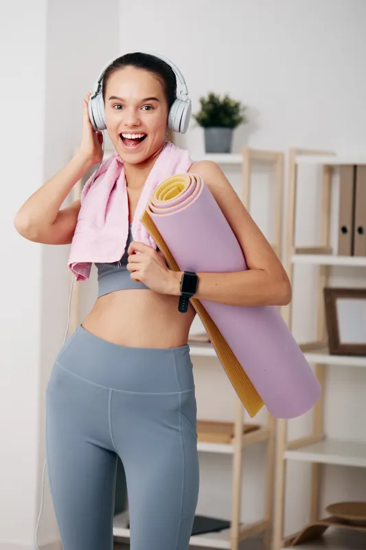 Fitness woman in a gray sports bra and blue leggings, wearing headphones, carrying a rolled yoga mat and towel in a bright home studio with shelves and a plant, ready for an energetic workout.