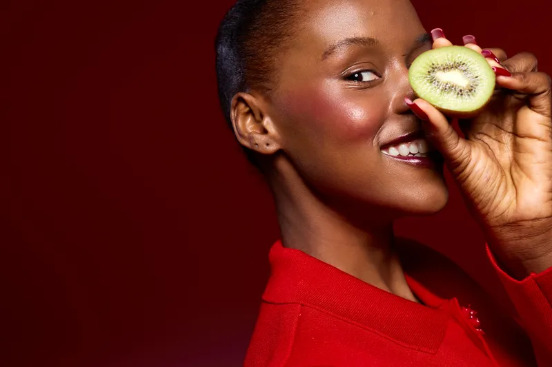 Smiling African American woman with dark skin holding half of kiwi fruit near her face on red background. Concept of healthy lifestyle, natural beauty, skincare, and fresh fruit nutrition.