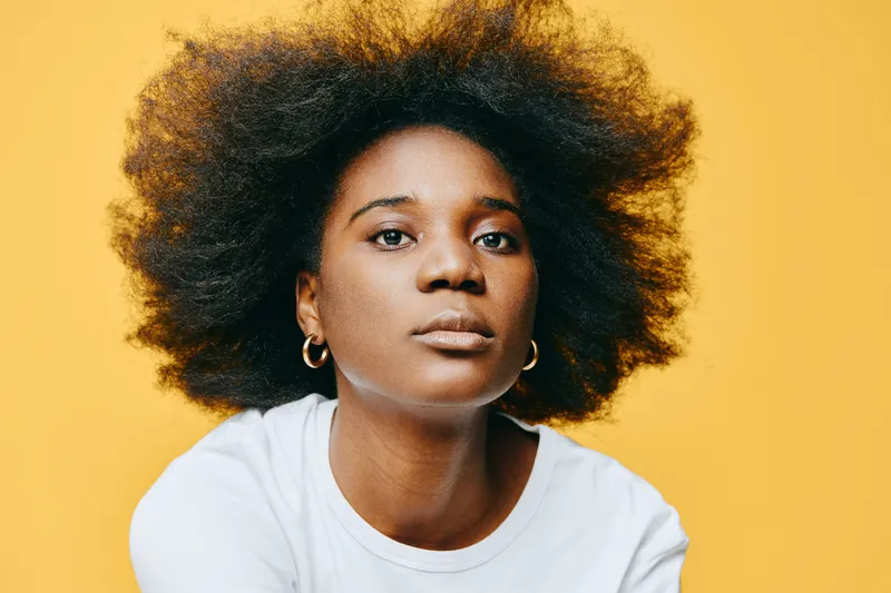 Young African woman with natural curly hair expressing confidence against a bright yellow background, wearing a simple white t shirt, ideal for wellness and beauty concepts