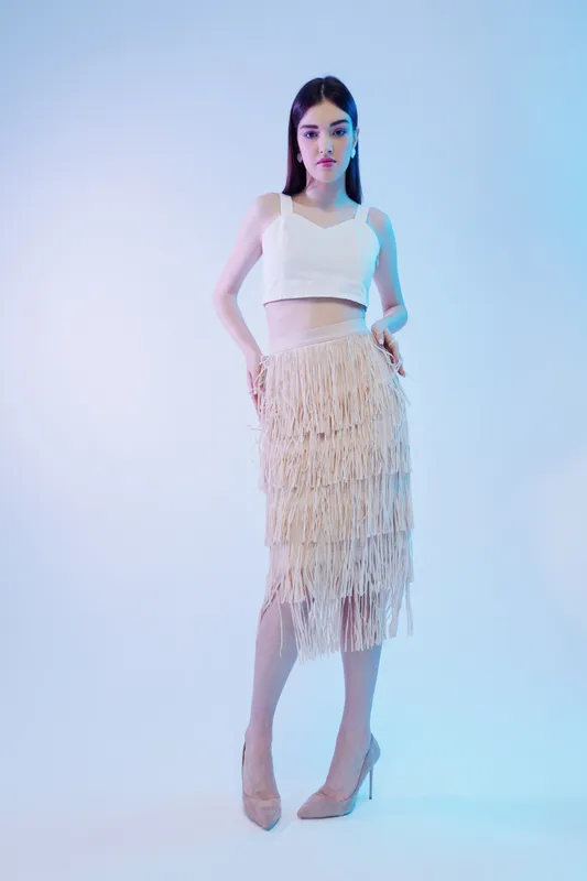 Young woman in a white crop top and beige fringed skirt poses confidently in high heels against a soft blue background. Concept of beauty fashion and style.