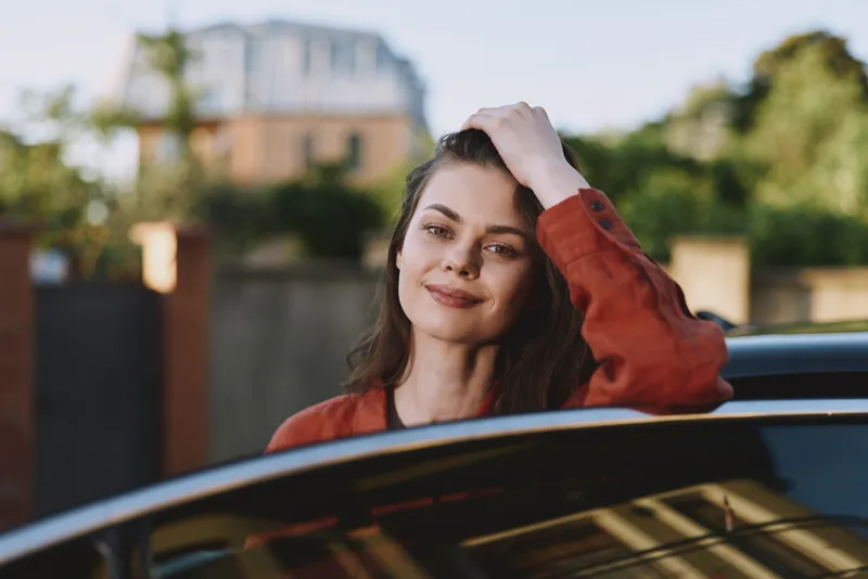 A smiling woman leans from a car window in daytime, portraying a casual lifestyle moment outdoors. Bright urban setting, relaxed pose, mood of freedom and confidence