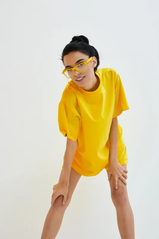 young woman in yellow oversized t-shirt and matching glasses posing confidently on plain white background with casual hairstyle and relaxed expression