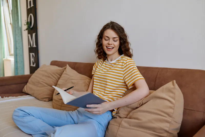 Smiling woman with curly hair sits on a sofa wearing a striped shirt, reading a notebook. Home interior, casual lifestyle, and self-care vibe. Soft lighting creates a warm, cozy mood.