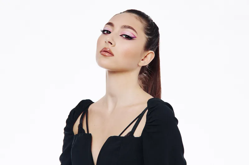 Confident woman with creative pink eye makeup and glossy lips poses against white background in trendy black outfit. Concept for beauty fashion beauty editorial photography.