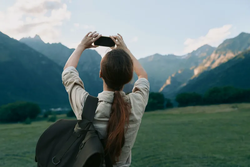 Woman with backpack taking a photo in a mountainous landscape, exploring outdoors. Bright natural light highlights nature, travel, and everyday wellness moments.