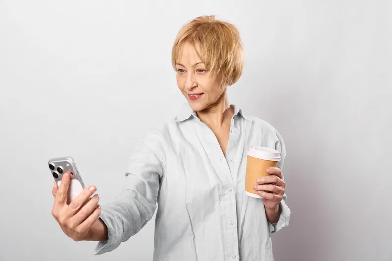 Senior woman with smartphone payment using credit card for online shopping, holding mug and phone, calm focused expression, studio shot on white background