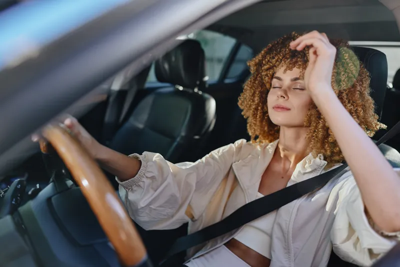 Relaxed woman with curly hair driving car, wearing casual light jacket and seatbelt, enjoying sunlight with eyes closed inside modern vehicle interior.