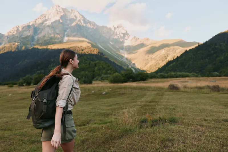 A woman hiker with a backpack stands in an open meadow, facing distant mountains, capturing a moment of outdoor adventure and solo nature travel. In bright daytime scenery.