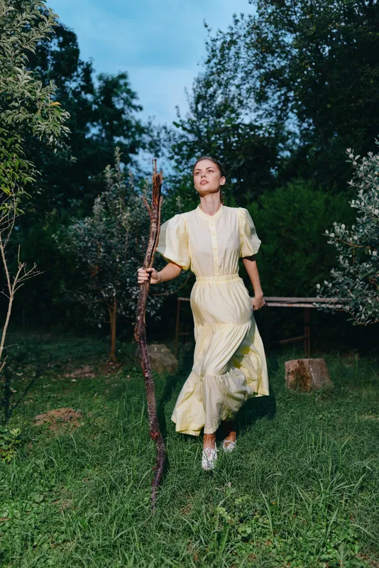 Woman walking in nature with wooden staff and long yellow dress, portrait with authenticity inclusivity focus, meadow scene and calm outdoor mood