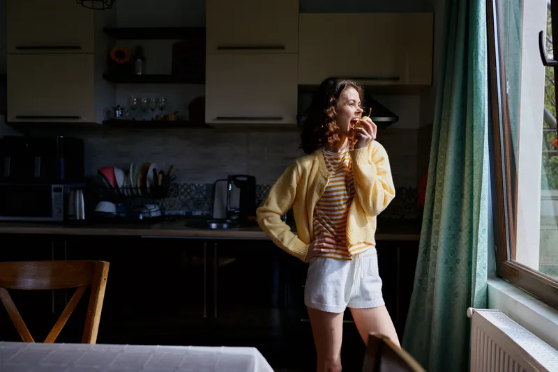 Woman in a yellow cardigan and striped top stands by a sunlit window in a cozy kitchen, enjoying a snack and a relaxed casual home moment, captured candidly.
