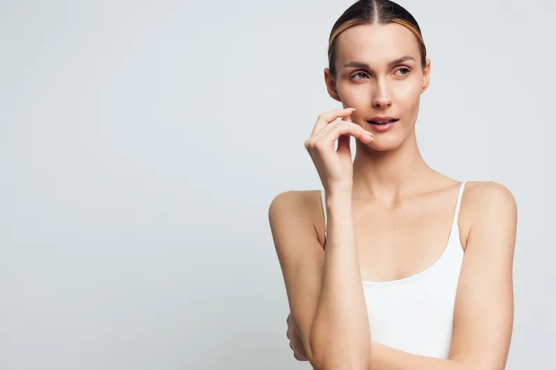 Young woman studio portrait holding smartphone to ear making a phone call, calm confident communication, white tank top, looking at camera, soft light gray background, close up
