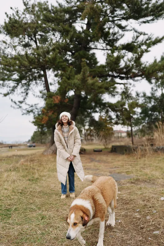 Woman and dog outdoors in a quiet field, wearing a warm coat and knit hat. Calm rural scene with open sky, natural light and a relaxed, friendly mood.