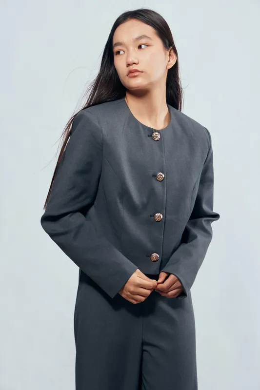 Young woman in gray tailored suit with gold buttons posing with hands clasped looking sideways on light background. Elegant fashion, casual style and professional appearance portrait.