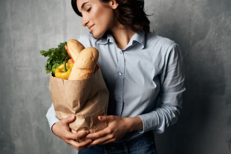 Весёлая брюнетка с пакетом продуктов, в голубой рубашке, на фоне овощей и тёмного фона