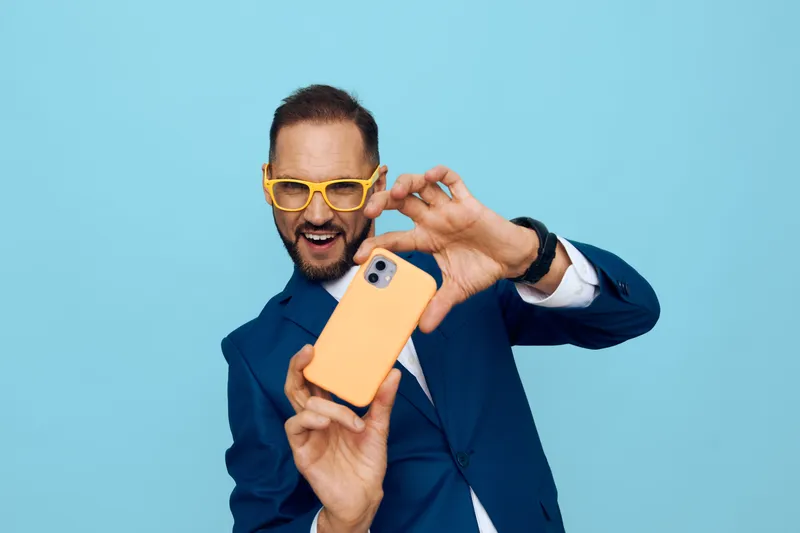 A confident business professional poses against a vibrant blue backdrop, taking a playful selfie with a smartphone, showcasing enthusiasm, focus, and approachability.