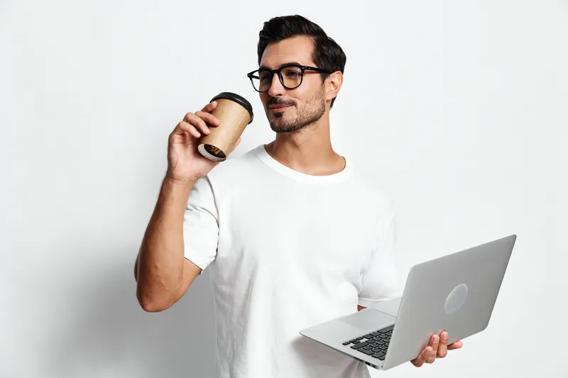 Confident young man with glasses holding coffee cup while using laptop, smiling and looking away. Modern workspace concept against plain background.