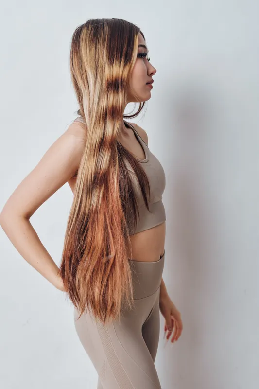 Young woman with long straight hair standing sideways wearing beige sportswear indoors. Female model with casual pose against plain background showing natural hairstyle and fitness outfit.