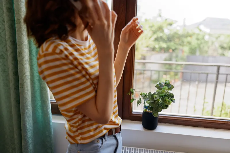 A woman in a striped shirt stands by a window, looking outside with her hand near her face in a bright home setting, soft morning light, plant on the sill, casual home environment.